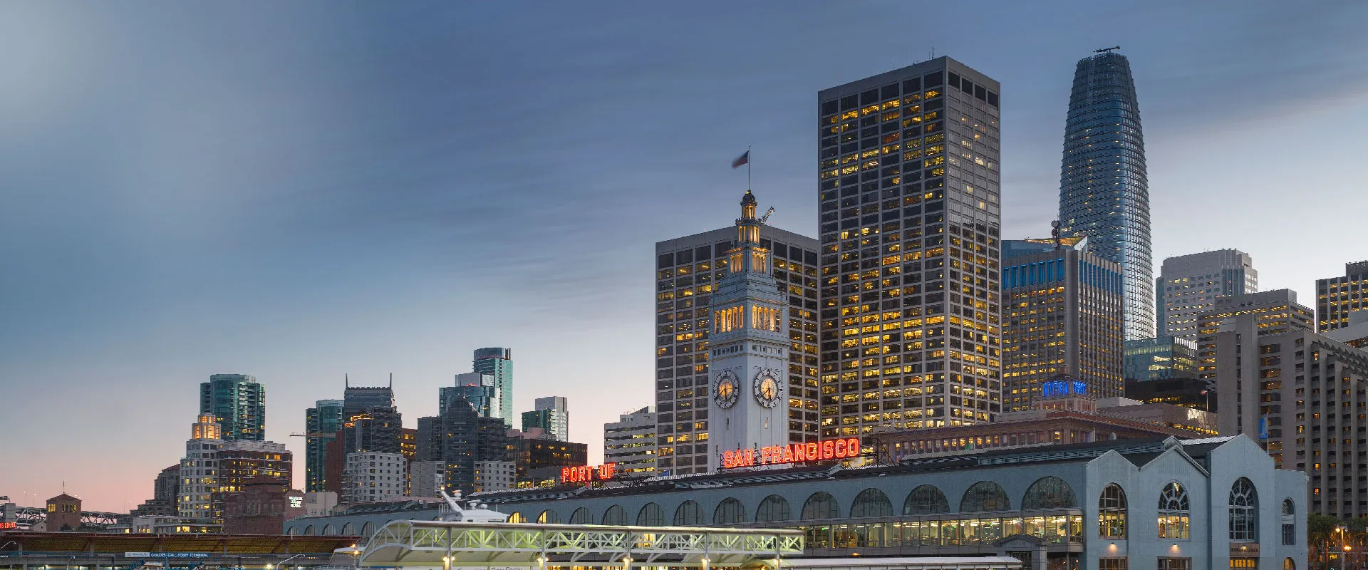 San Francisco Ferry Building clock tower at dusk, with lit downtown office towers and Salesforce Tower behind